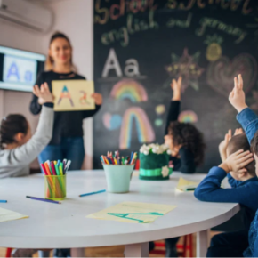 a teacher holding up a letter A to a group of students raising their hands. 